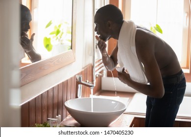 Happy smiling african american guy with towel on shoulders bending over sink with running water in modern bathroom at home, washing face, looking in mirror. Daily personal morning hygiene routine. - Powered by Shutterstock