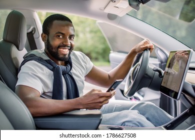 Happy Smiling African American Male Driver Sitting Behind The Self-driving Steering Wheel Of An Autonomous Electric Modern Car. Happy Guy Holds Phone And Smiles To Camera In Modern Electric Car