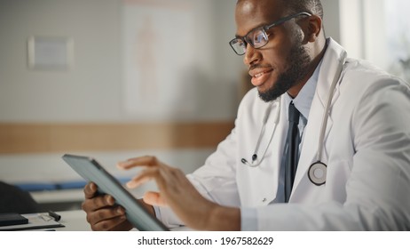 Happy and Smiling African American Male Doctor Wearing White Coat Working on Tablet Computer at His Office. Medical Health Care Professional Working with Test Results, Patient Treatment Planning.