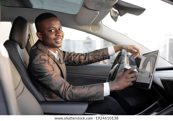 Happy smiling African American male driver sitting\
behind the self-driving steering wheel of an autonomous electric\
modern car. Happy guy holds coffee cup and smiles to camera in\
modern electric car