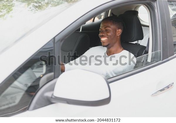 Happy smiling African American male driver sitting
behind the self-driving steering wheel of an autonomous electric
modern car. Happy guy holds phone and smiles to camera in modern
electric car