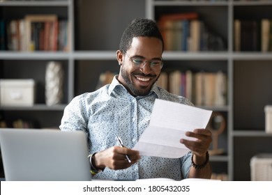 Happy smiling African American man wearing glasses reading letter at workplace, sitting at work desk, satisfied businessman received good news, job promotion, money refund or great exam results