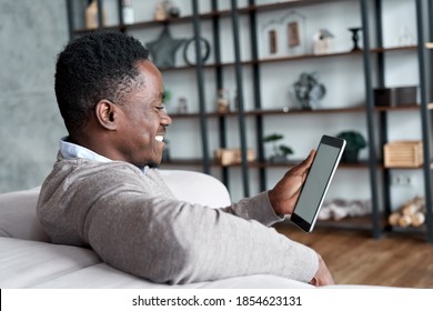 Happy Smiling African American Man Holding Digital Tablet Computer Sitting On Couch At Home. Young Adult Black Guy Using Apps On Tab Technology Device Watching Videos Or Browsing Internet.