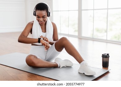 Happy Smiling African American Sportswoman Using Smartwatch Fitness Tracker After Workout, Checking Burned Calories, Setting Wristwatch, Wearing Wireless Heaset For Music Sitting On Floor Mat Indoors - Powered by Shutterstock