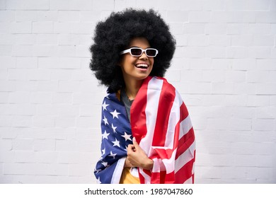 Happy Smiling African American Teenager Wearing Sunglasses Wrapped In Usa Flag Celebrating July 4th Forth Independence Day Of United States. Patriotic Portrait On White Bricks Wall Background.