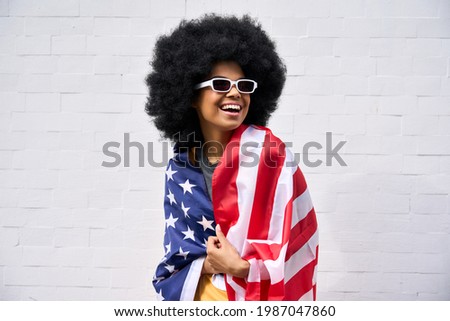 Happy smiling African American teenager wearing sunglasses wrapped in usa flag celebrating july 4th forth independence day of united states. Patriotic portrait on white bricks wall background.