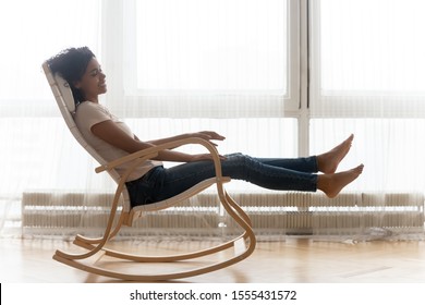 Happy smiling African American woman relaxing in comfortable wooden rocking chair at home, satisfied girl resting in cozy armchair, spending lazy day weekend indoors, daydreaming, enjoying free time