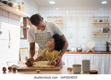 Happy Smiling African Family  In Aprons Cooking And Kneading Dough On Wooden Table. Black Father With Son In Yellow Shirt Baking Cake Together And Looking At Each Other In White Home Kitchen Interior.