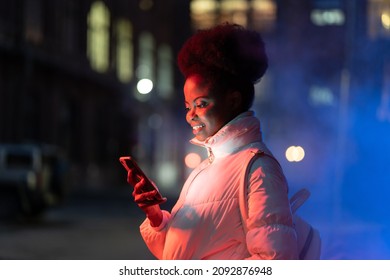 Happy Smiling African Girl Hold Smartphone Looking At Screen And Reading Message While Waiting For Friends In City At Night. Black Woman With Cellphone Outdoors Texting Scrolling Social Media Online