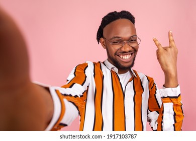 Happy Smiling African Man With Dreadlocks In Stylish Striped Shirt Showing Rock And Roll Gesture With Fingers, Posing At Selfie Camera, Live Stream. Indoor Studio Shot Isolated On Pink Background