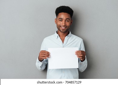 Happy Smiling African Man Holding Blank Sheet Of Paper While Standing Isolated Over Gray Background