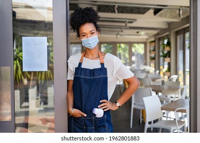 Happy Smiling African Woman Wearing Face Mask And Apron, Back To Work After Quarantine Due To Covid19. Portrait Of Black Waitress Showing Sign No Mask No Entry In Her Small Business Shop After Covid.