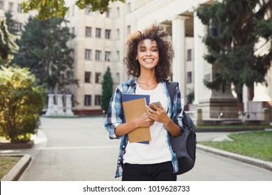 Happy Smiling African-american Student Girl With Backpack At University Background. Woman With Mobile And Workbooks Having Rest In Campus During Lunch Break. Technology, Education, Leisure Concept