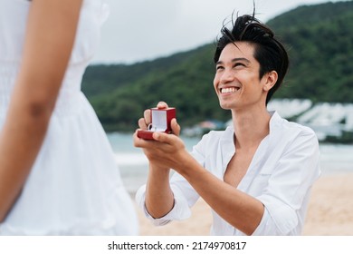 Happy Smiling Asian 30s Man Proposing Woman With A Wedding Ring On The Beach In Summer. A Couple Wearing A White Shirt And Dress - Relationship Goals And Marriage Proposal Concept