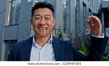 Happy smiling Asian adult 40s man holding keys portrait outdoors win reaction victory celebrate male realtor real estate agent investor buyer selling property new building office dwelling skyscrapers