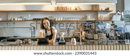 Happy smiling asian barista, girl behind counter, working with POS terminal and brewing filter kit, making coffee in cafe.