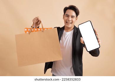 Happy smiling Asian businessman holding an empty white smart phone screen and a lot of shopping bags on the other side Fashionable joyful man in white shirt and suit on beige color background. - Shutterstock ID 2258352833