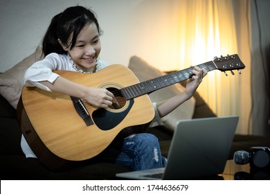 Happy smiling asian child girl is singing,playing the guitar to enjoy,fun,student is studying tutor with teacher,video conference calling on laptop computer,learn teach in online,distance teaching 