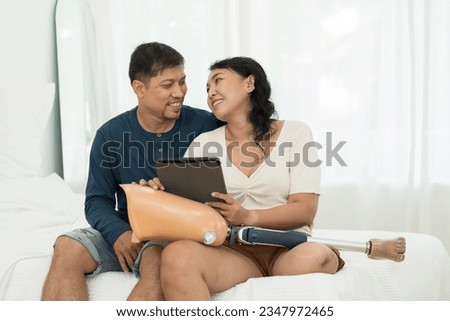 Happy smiling Asian couple using digital tablet and holding prosthetic leg sitting in bedroom at home. Asian woman with prosthetic leg spending time with her husband at home