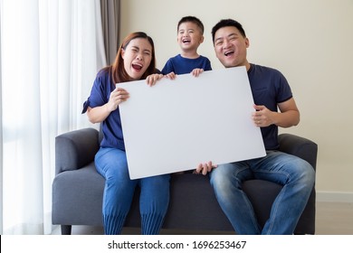 Happy Smiling Asian Family Holding Blank Big White Poster And Sitting On Sofa In Living Room, Wow And Surprised Concept