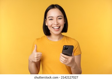 Happy Smiling Asian Girl Holding Mobile Phone And Showing Thumbs Up, Recommending Application On Smartphone, Standing Over Yellow Background