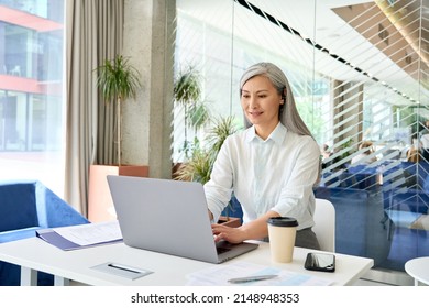 Happy Smiling Asian Older Businesswoman Executive Manager In Headset Sitting At Desk Working Typing On Pc Laptop Consulting Customer On Computer In Contemporary Office. Business Technologies Concept.