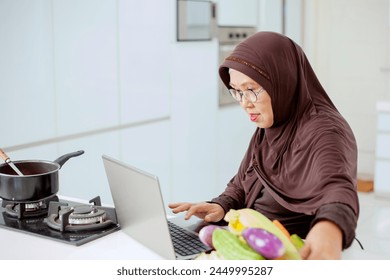 Happy smiling asian older muslim woman looking at a laptop computer in apron cooking healthy food - Powered by Shutterstock