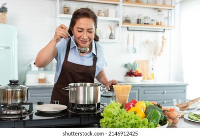 Happy smiling asian older woman in apron is tasting and smell food while cooking in kitchen. - Shutterstock ID 2146912425