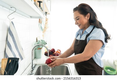 Happy Smiling Asian Older Woman In Apron Washing The Apple In Kitchen. Housewife Cooking Concept.