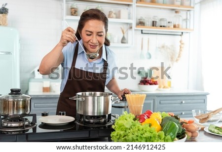Happy smiling asian older woman in apron is tasting and smell food while cooking in kitchen.