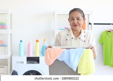 Happy Smiling Asian Senior Elderly Woman Housewife Carrying Clothes Basket For Doing Laundry At Laundry Room With Washing Machine, Grandma Doing Housework And Cleaning Clothes At House.