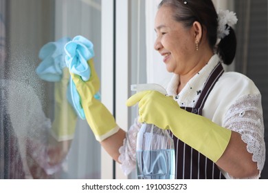 Happy Smiling Asian Senior Elderly Woman Housewife Wearing Rubber Gloves And Holding Cleaning Spray And Clothe For Wiping Window, Cleaning Up House, Grandma Doing Housework And Wipeing The Glass.