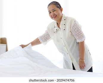 Happy Smiling Asian Senior Elderly Woman Housewife Cleaning And Making Bed At Bedroom, Grandma Doing Housework And Cleaning, Old Female Chambermaid Changing The White Blanket And Sheet.