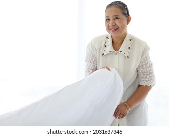 Happy Smiling Asian Senior Elderly Woman Housewife Cleaning And Making Bed At Bedroom, Grandma Doing Housework And Cleaning, Old Female Chambermaid Changing The White Blanket And Sheet.