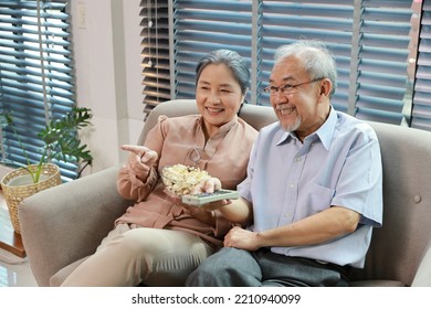 Happy Smiling Asian Senior Man And Woman Sitting On Sofa And Eating Popcorn While Having Fun With Movie Rest Indoor At Home Living Room. Couple Elder Husband And Wife Embrace Are Happy While Watch TV