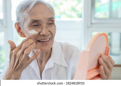 Happy smiling asian senior woman apply sunscreen lotion on the cheek,protect her beautiful face,old elderly using skin care product for sensitive skin,sun block day cream on facial skin in sunny day
