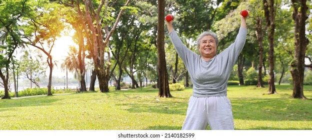 Happy And Smiling Asian Senior Woman Doing Arm Work Out And Lifting Dumbbell Exercise With Relaxation For Healthy In Park Outdoor After Retirement. Health Care Elderly Outdoor Lifestyle Concept.