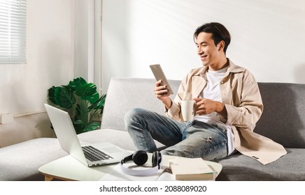 Happy smiling Asian student sits and relaxes on a couch using modern tablet browsing unlimited wireless internet, positive young man freelancer working uses wifi at a home apartment, technology idea. - Shutterstock ID 2088089020