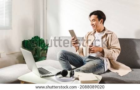Happy smiling Asian student sits and relaxes on a couch using modern tablet browsing unlimited wireless internet, positive young man freelancer working uses wifi at a home apartment, technology idea.