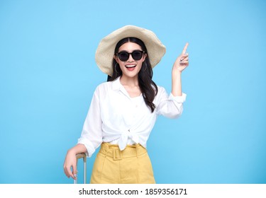 Happy smiling Asian woman dressed in summer clothes and wearing hat with luggage enjoying their summer vacation  and pointing finger a copy space on bright blue background.