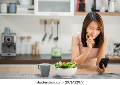 Happy smiling Asian woman using smartphone mobile at home in kitchen with salad bowl on the table - Powered by Shutterstock