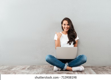 Happy smiling asian woman working on laptop computer while sitting on the floor with legs crossed and pointing finger away isolated over gray background