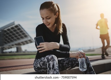 Happy Smiling Athlete Woman Using Smartphone Device In Case Holder On Her Arm, Attractive Fitness Girl Checking Her Progress On Touchscreen Of Cellphone While Resting After Workout, Flare Light