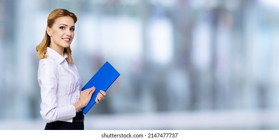 Happy Smiling Attractive Businesswoman, With Copy Space Area For Some Slogan Or Text. Confident Blond Woman In White Shirt With Blue Folder, Over Blurred Office Background. Blurred Office Background.