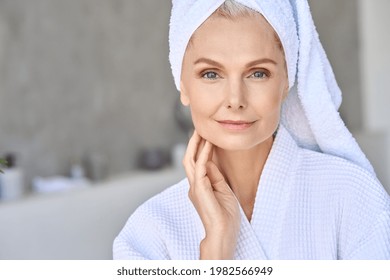 Happy Smiling Attractive Senior Older Woman Wearing Bathrobe And White Towel Touching Face Looking At Camera At Bath Room. Advertising Of Skin Care Spa Procedures Concept. Closeup Portrait.