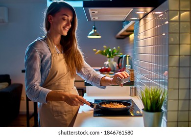 Happy Smiling Attractive Young Cooking Woman Housewife Salting And Preparing Food In A Frying Pan On The Stove For Evening Dinner At Modern Loft Style Kitchen                      