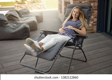 Happy Smiling Attractive Young Pregnant Young Woman Relaxing Outdoors On A Recliner Chair On A Wooden Deck Cradling Her Baby Bump In Her Hands