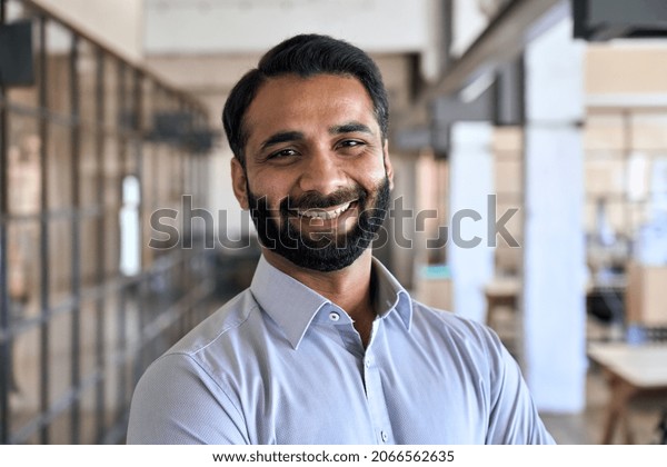 Happy smiling bearded indian business man small\
business owner, company leader or sales manager, male hispanic ceo\
executive, successful lawyer looking at camera standing in office,\
headshot portrait.