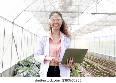 Happy smiling beautiful Asian botanist scientist woman in lab coat holding laptop computer, female biological researcher working on experimental plant plots in greenhouse. Biology Agricultural Science