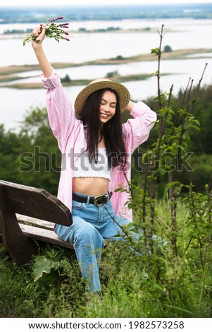 Happy smiling beautiful girl in a straw boater hat sits on a bench with a picturesque view with a bouquet of flowers.